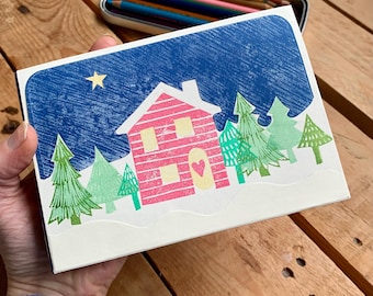 May include: A rectangular greeting card featuring a block-print style illustration of a pink house with a heart on the door, surrounded by green pine trees against a dark blue night sky with a single yellow star, set on a white base.