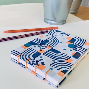 May include: A rectangular notebook with exposed orange thread binding and a white cover featuring a repeating pattern of dark blue arches, solid orange squares, and clusters of small dark blue dots, accompanied by two colored pencils.