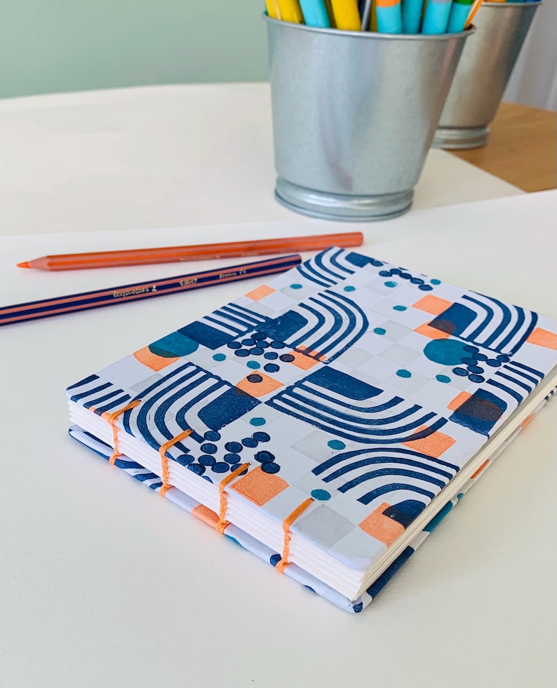 May include: A rectangular notebook with exposed orange thread binding and a white cover featuring a repeating pattern of dark blue arches, solid orange squares, and clusters of small dark blue dots, accompanied by two colored pencils.