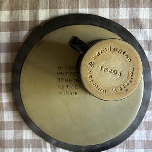 May include: A ceramic saucer and mug set with a dark brown rim and a light tan center. The saucer features the stamped text BENNINGTON POTTERS BENNINGTON VERMONT 1669. The mug base displays the text bennington potters vermont 1626.