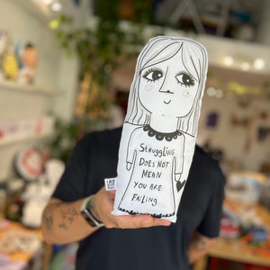 Könnte beinhalten: Weiße Puppe mit einer Schwarz-Weiß-Illustration eines Frauen-Gesichts und -Körpers. Die Puppe hat lange Haare und ein Kleid. Auf der Puppe steht der Text "Struggling Does Not Mean You Are Failing."