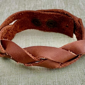 May include: A brown leather bracelet featuring a braided design. The bracelet is constructed from a single strip of leather that is looped and twisted to create a woven appearance, secured at the back with two dark metal snap button fasteners.