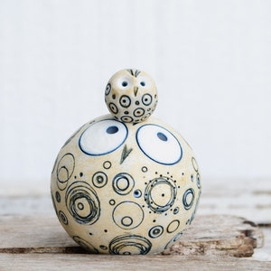 May include: A ceramic owl sculpture with a white and yellow speckled pattern and blue accents. The owl has large, white eyes and a smaller owl perched on top of its head.