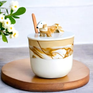 Puede incluir: Una taza de cerámica blanca con un diseño marmoleado marrón y blanco. La taza tiene una tapa con una figurita de dachshund en la parte superior. La taza está sobre un posavasos de madera.