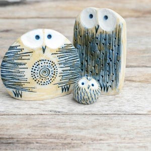 Könnte beinhalten: Drei Keramik-Eulenfiguren in verschiedenen Größen. Die Eulen haben eine cremefarbene Basis mit blauen und schwarzen Details. Die größte Eule ist rechteckig, die mittlere Eule ist oval und die kleinste ist rund.