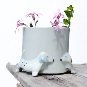 May include: A white ceramic planter shaped like a dachshund with pink flowers inside. The planter has blue and yellow polka dots on the side.