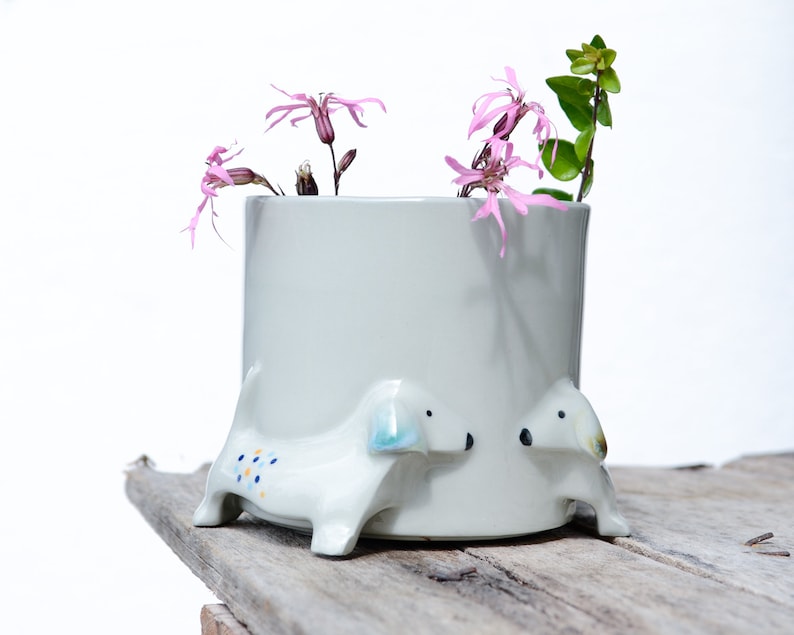 May include: A white ceramic planter shaped like a dachshund with pink flowers inside. The planter has blue and yellow polka dots on the side.