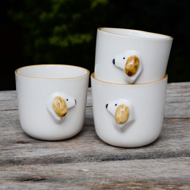 May include: Three white ceramic mugs with a brown dog head design. The mugs are stacked on top of each other on a wooden surface.