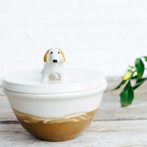May include: A white ceramic bowl with a lid, featuring a small dog figurine on top. The bottom half of the bowl has a brown, marbled design. The dog has a white body with tan ears. The bowl is likely for kitchen or home decor.