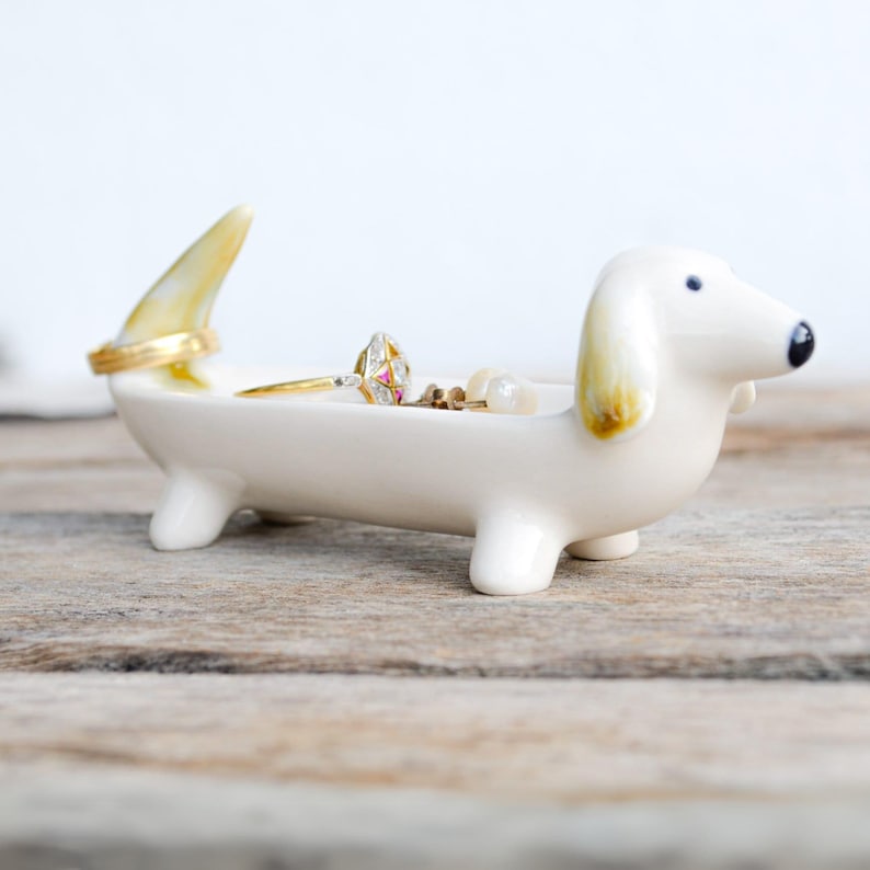 Pode incluir: Porta-an&eacute;is de cer&acirc;mica branca em forma de dachshund com um anel de ouro e um pequeno anel com pedras preciosas dentro. O dachshund tem orelhas amarelas e est&aacute; sentado em uma superf&iacute;cie de madeira.