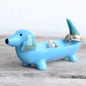 Pode incluir: Um prato de cer&acirc;mica azul claro em forma de dachshund com a cauda ereta. O prato cont&eacute;m alguns an&eacute;is e brincos.