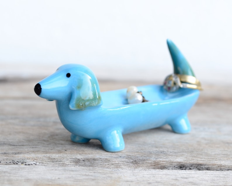 Pode incluir: Um prato de cer&acirc;mica azul claro em forma de dachshund com a cauda ereta. O prato cont&eacute;m alguns an&eacute;is e brincos.