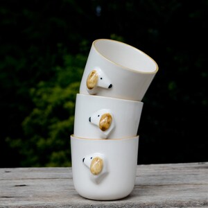 May include: Three white ceramic mugs stacked on top of each other. Each mug has a small, brown and white dog face sculpted on the side.