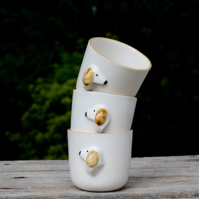 May include: Three white ceramic mugs stacked on top of each other. Each mug has a small, brown and white dog face sculpted on the side.
