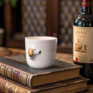 May include: White ceramic mug with a 3D dog head design. The dog has brown ears and a black nose. The mug is sitting on a stack of books.