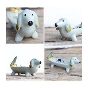 Pode incluir: Um porta-an&eacute;is de cer&acirc;mica em forma de dachshund com um esmalte azul claro e detalhes dourados. O dachshund est&aacute; deitado com a cabe&ccedil;a virada para o lado. O porta-an&eacute;is tem um pequeno prato para an&eacute;is e outras joias.