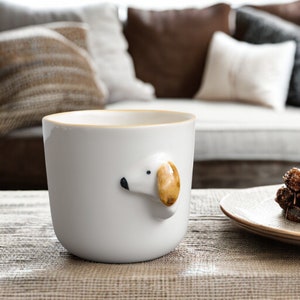 May include: White ceramic mug with a brown and white dog head sticking out of the side. The mug is sitting on a brown woven surface.
