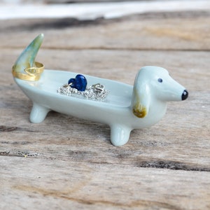 Pode incluir: Um prato de cer&acirc;mica azul claro em forma de dachshund com um anel dourado e contas de vidro azuis. O prato est&aacute; sobre uma superf&iacute;cie de madeira.