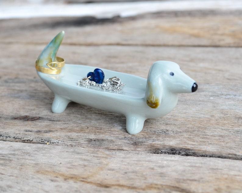 Pode incluir: Um prato de cer&acirc;mica azul claro em forma de dachshund com um anel dourado e contas de vidro azuis. O prato est&aacute; sobre uma superf&iacute;cie de madeira.