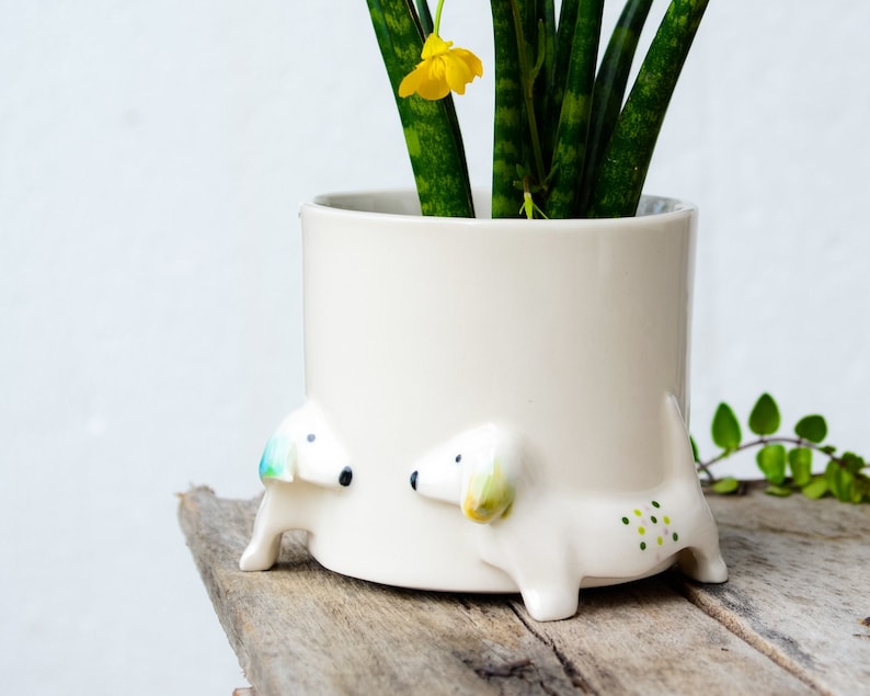 May include: White ceramic planter with two small dachshund figurines on the bottom. The planter has a green plant with a yellow flower inside.