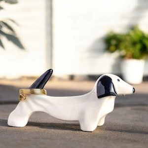 May include: White ceramic dachshund ring holder with black ears and tail. The dachshund is lying down with its head turned to the side. A gold ring is placed on the tail.