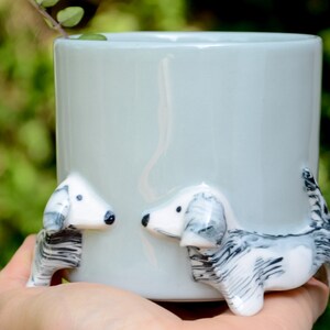 May include: A light blue ceramic mug with two white and black dachshund figurines attached to the sides. The mug is held in a hand.