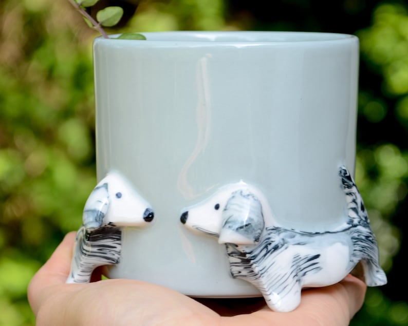 May include: A light blue ceramic mug with two white and black dachshund figurines attached to the sides. The mug is held in a hand.