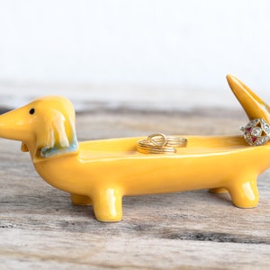 Pode incluir: Um porta-an&eacute;is de cer&acirc;mica amarelo em forma de dachshund com tr&ecirc;s an&eacute;is dourados no topo. O dachshund est&aacute; em p&eacute; sobre uma superf&iacute;cie de madeira.