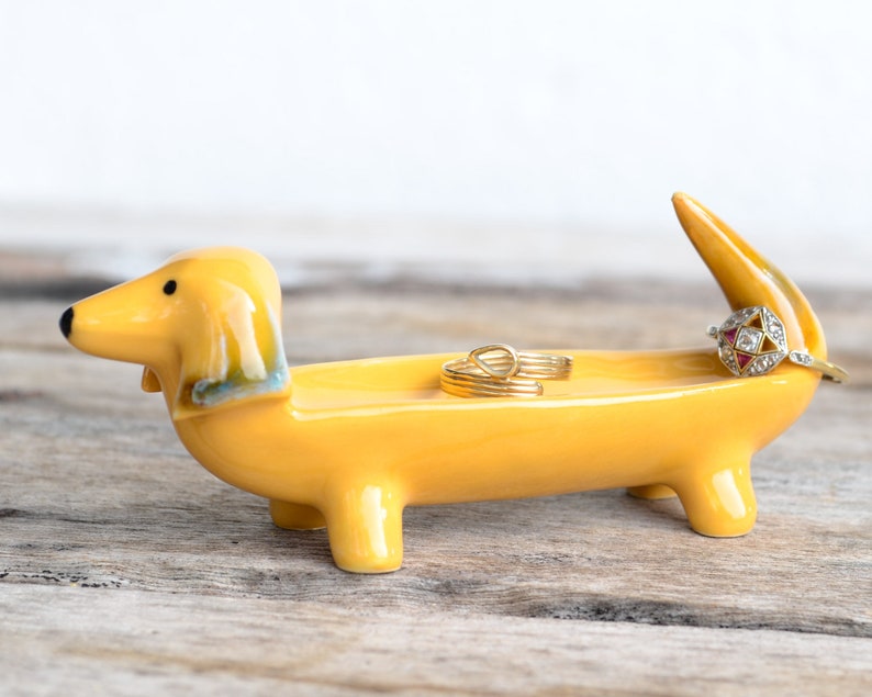 Pode incluir: Um porta-an&eacute;is de cer&acirc;mica amarelo em forma de dachshund com tr&ecirc;s an&eacute;is dourados no topo. O dachshund est&aacute; em p&eacute; sobre uma superf&iacute;cie de madeira.