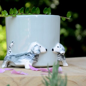 May include: A light blue ceramic planter with two white and gray ceramic dachshund dogs on the sides. The dogs are facing each other and have black eyes and noses.