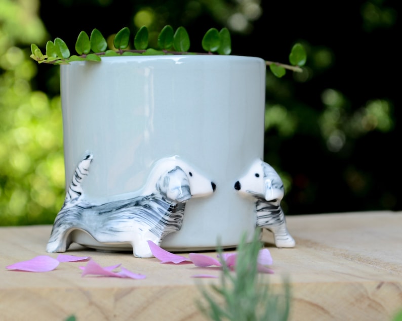 May include: A light blue ceramic planter with two white and gray ceramic dachshund dogs on the sides. The dogs are facing each other and have black eyes and noses.