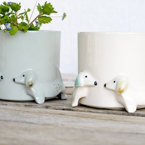 May include: Two ceramic planters shaped like dogs, one is white and the other is light gray. Both planters have small green and blue dots on the sides. The planters are on a wooden surface.