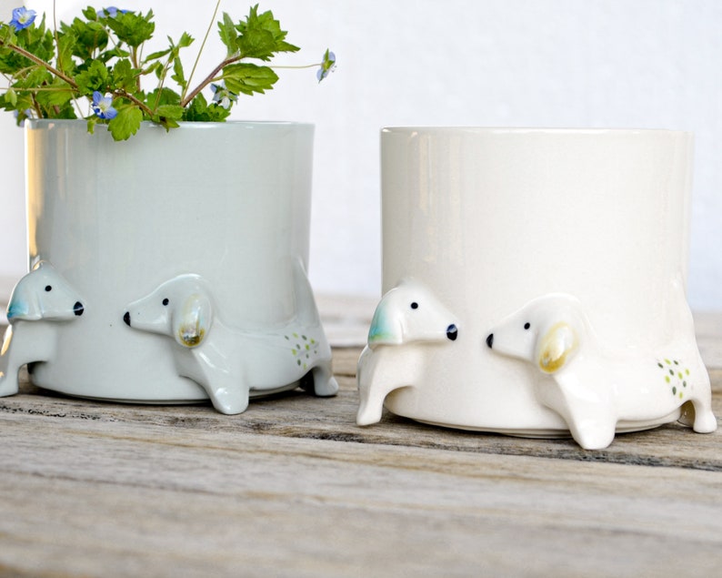 May include: Two ceramic planters shaped like dogs, one is white and the other is light gray. Both planters have small green and blue dots on the sides. The planters are on a wooden surface.