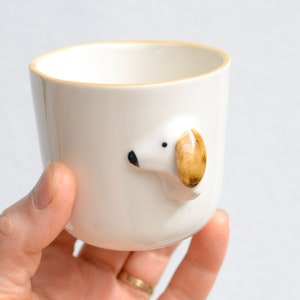 May include: White ceramic mug with a brown and white dog head sticking out of the side. The dog's ear is brown and the rest of the dog is white.