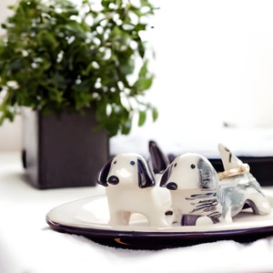 May include: Two ceramic dog figurines on a white and black dish. The dogs are white with black markings and have black eyes. One dog is sitting up and the other is lying down.