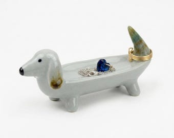 May include: A light grey ceramic dachshund-shaped jewellery tray with a smooth finish. The dog's tail is an upright, mottled green and brown stone-like accent. A silver chain with a blue heart pendant and a gold ring rest inside the tray.