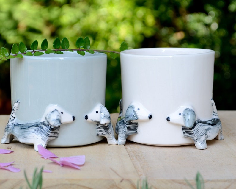 May include: Two ceramic planters shaped like mugs with dachshund dog figurines on the bottom. One planter is light blue and the other is white.