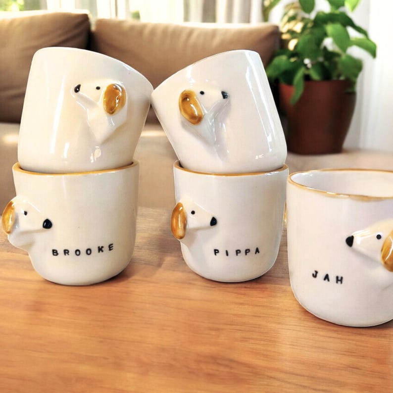 May include: Four white ceramic mugs with dog faces and names on the front. The mugs are Brooke, Pippa, and Jah.