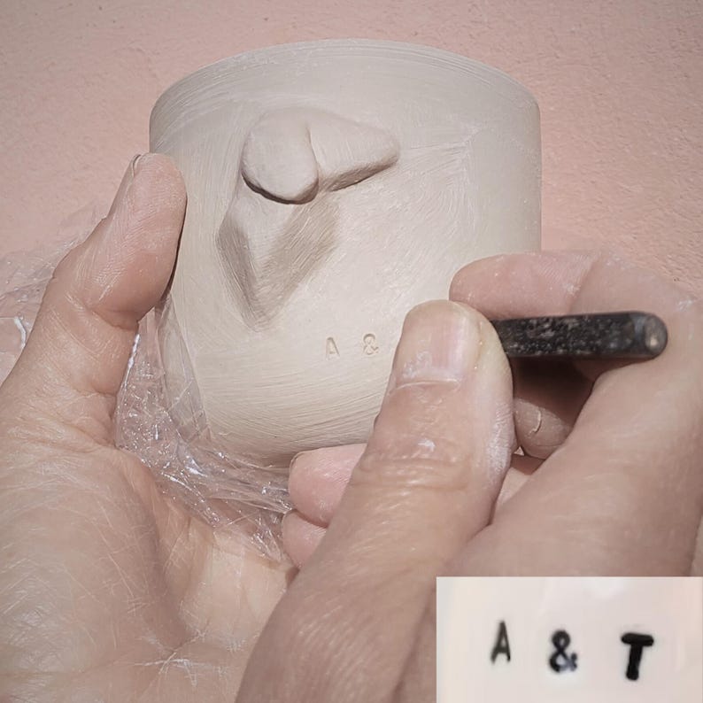 May include: A close-up of a person's hands stamping the letters "A &" into a clay cup. The cup has a raised, textured design on the side.