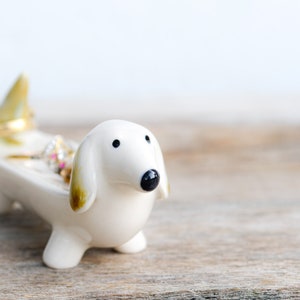 Peut inclure: Porte-bagues en céramique blanche en forme de dachshund avec des anneaux dorés et argentés à l'intérieur. Le porte-bagues est posé sur une surface en bois.