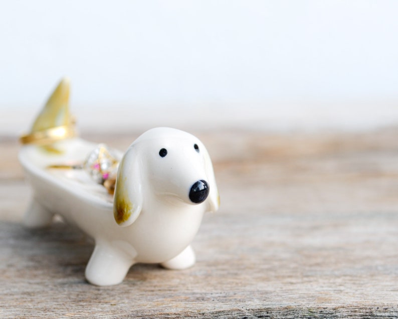 Pode incluir: Porta-an&eacute;is de cer&acirc;mica branca em forma de dachshund com an&eacute;is de ouro e prata dentro. O porta-an&eacute;is est&aacute; sobre uma superf&iacute;cie de madeira.