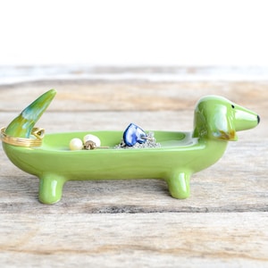 Pode incluir: Prato de cer&acirc;mica verde em forma de dachshund com um anel de ouro, brincos de p&eacute;rola e um pingente de cora&ccedil;&atilde;o azul dentro.