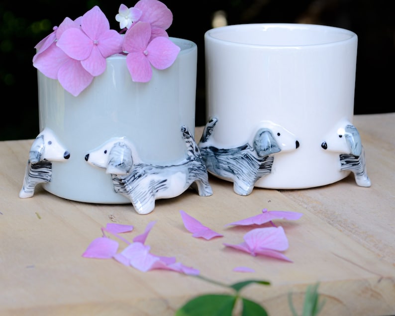 May include: Two ceramic planters with dachshund dog figurines on the sides. The planters are white and light blue. The planter on the left has pink flowers in it.
