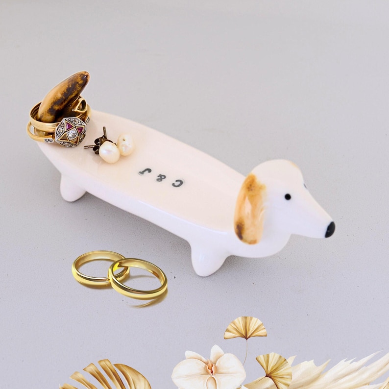 Pode incluir: Prato de cer&acirc;mica branca em forma de dachshund com orelhas marrons. O prato tem as iniciais "C & J" escritas nele. H&aacute; an&eacute;is e brincos no prato.