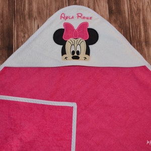 Op de afbeelding: Roze badcape met witte bies en een wit geborduurd gezicht van Minnie Mouse met een roze strik. De badcape heeft de naam "Apla Rose" geborduurd op de capuchon.