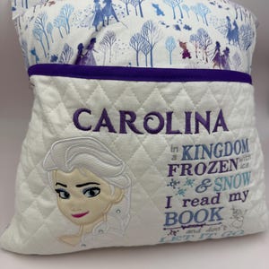 Op de afbeelding: Een wit gewatteerd kussen met de naam "CAROLINA" in paars geborduurd. Het heeft een Elsa-applicatie en de tekst "In a KINGDOM FROZEN with ice & SNOW I read my BOOK and don't LET IT GO."