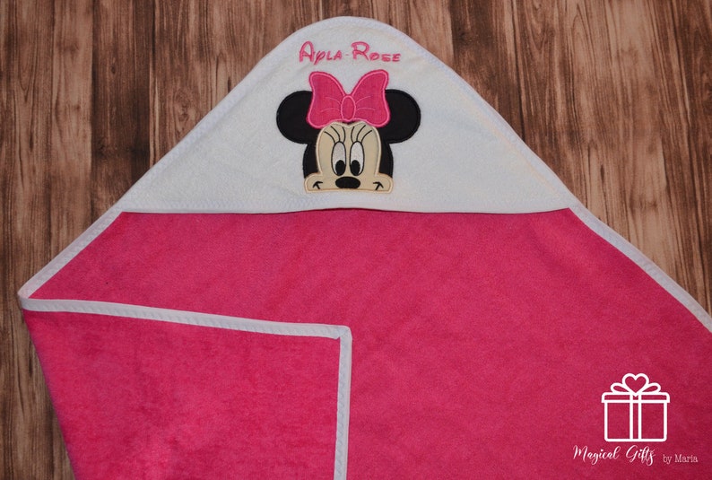 Op de afbeelding: Roze badcape met witte bies en een wit geborduurd gezicht van Minnie Mouse met een roze strik. De badcape heeft de naam "Apla Rose" geborduurd op de capuchon.