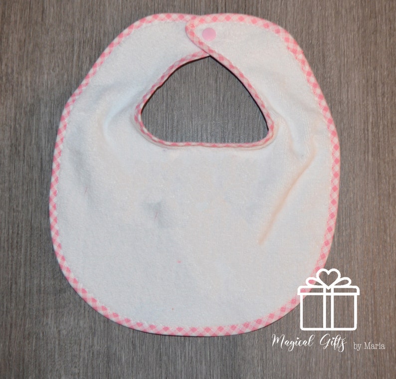 Op de afbeelding: Witte baby-slabbetje met een roze en witte geruite rand. Het slabbetje heeft een drukknoop sluiting aan de hals.