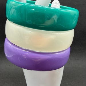 Könnte beinhalten: Drei gestapelte, klobige Armreifen in Türkis, Creme und Lila. Die Armbänder werden auf einer weißen, handförmigen Form vor einem dunklen Hintergrund präsentiert. Die Armreifen haben ein glänzendes Finish.