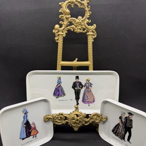 Könnte beinhalten: Ein Set aus drei weißen Keramiktabletts mit Illustrationen von Menschen in traditioneller Kleidung. Die Tabletts sind rechteckig und zeigen farbenfrohe Bilder von Figuren in historischer Kleidung. Eine goldfarbene dekorative Staffelei befindet sich hinter den Tabletts.